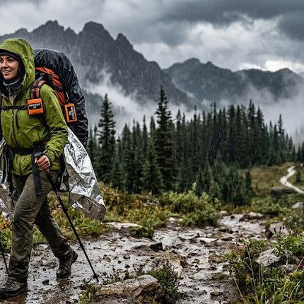 A hiker in a green jacket uses trekking poles on a rainy mountain trail. Her large backpack is partially covered by a reflective silver emergency poncho. Misty forests and jagged, stormy peaks create a rugged, high-stakes outdoor atmosphere.