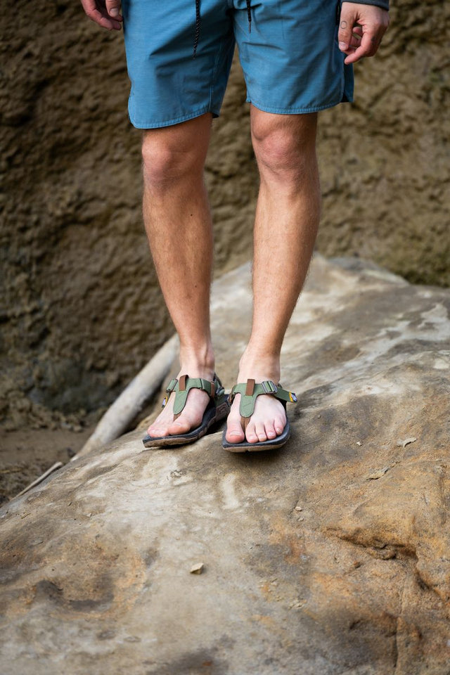 Man wearing Lems Switchback sandals while hiking on a rocky trail, stepping on a large rock with secure straps and rugged outsole visible in an outdoor setting.