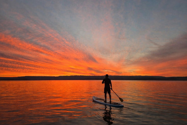 Person on stand-up-paddle board with an incredible beautiful orange sunset in the background on a smooth lake.
