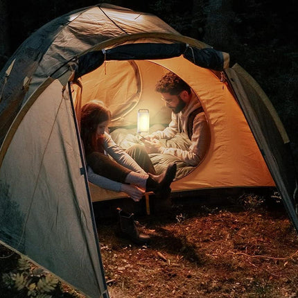 Man and woman sitting inside a tent in the woods at night, illuminated by a glowing lantern that casts warm light on their faces and surrounding camping gear.