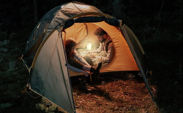 Man and woman sitting inside a tent in the woods at night, illuminated by a glowing lantern that casts warm light on their faces and surrounding camping gear.