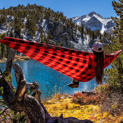 Woman sitting in a red Grand Trunk hammock suspended outdoors in front of a scenic lake and mountain landscape, relaxing in a peaceful natural setting.