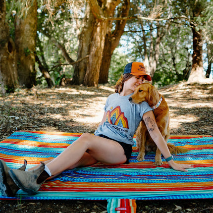 Woman sitting on a red, blue, yellow, and orange Grand Trunk Adventure Sheet outdoors with her dog, enjoying a relaxed camping scene in a natural setting.