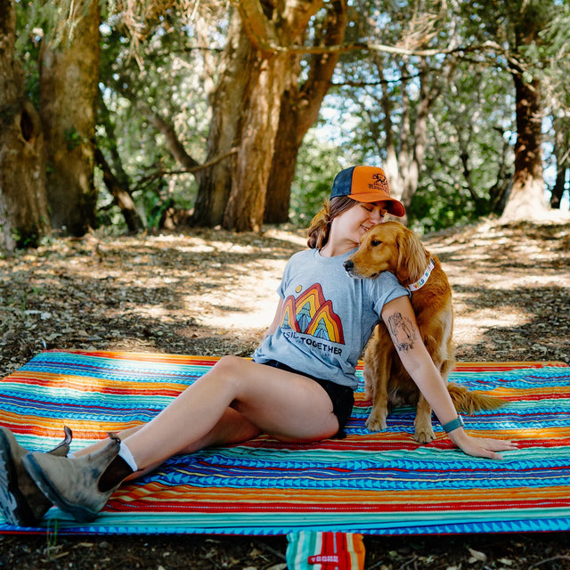 Woman sitting on a red, blue, yellow, and orange Grand Trunk Adventure Sheet outdoors with her dog, enjoying a relaxed camping scene in a natural setting.