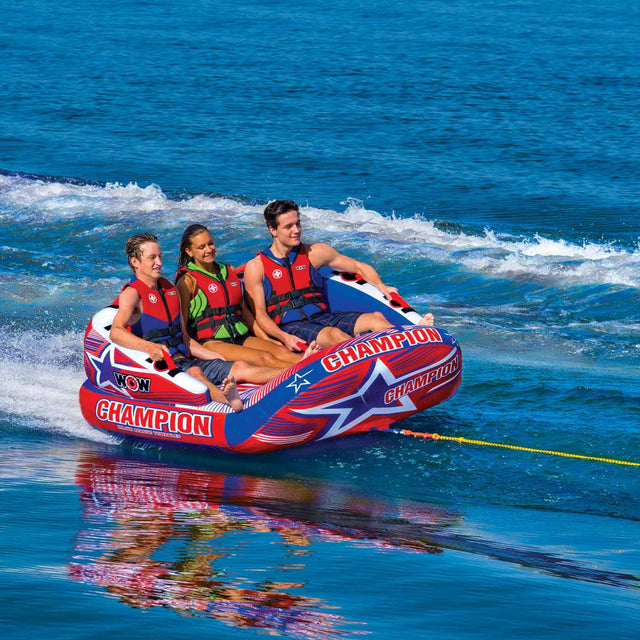 3 people riding a 3 person Wow Sports boating tube being pulled by a boat on a clear blue lake,