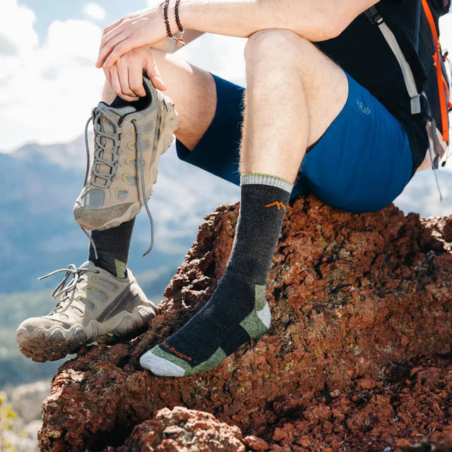 Hiker wearing Darn Tough crew hiking socks with merino wool cushioning, paired with trail shoes while resting on rocky mountain terrain outdoors.