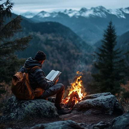 Man sitting on a rock at dusk reading a book by a campfire, surrounded by mountains and trees in a peaceful outdoor wilderness setting.