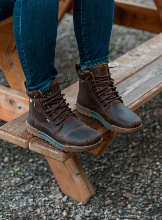 Person wearing dark wash jeans and brown leather boots with grey soles, sitting on a wooden bench