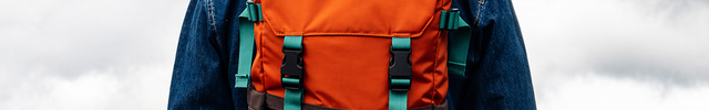 Man wearing a yellow beanie, denim shirt, and orange and brown backpack with teal straps, facing away