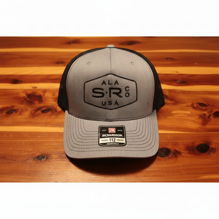 A Richardson 112 trucker hat in heather gray and black, featuring a black embroidered 'ALA SR Co USA' logo on the front, resting on a wooden surface.