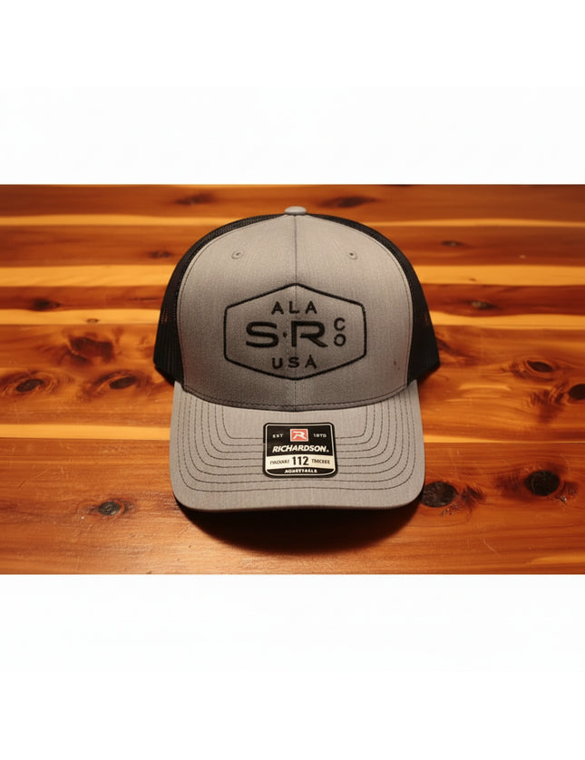 A Richardson 112 trucker hat in heather gray and black, featuring a black embroidered 'ALA SR Co USA' logo on the front, resting on a wooden surface.