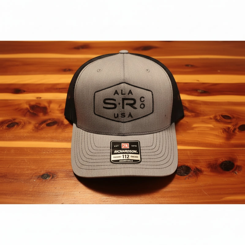 A Richardson 112 trucker hat in heather gray and black, featuring a black embroidered 'ALA SR Co USA' logo on the front, resting on a wooden surface.