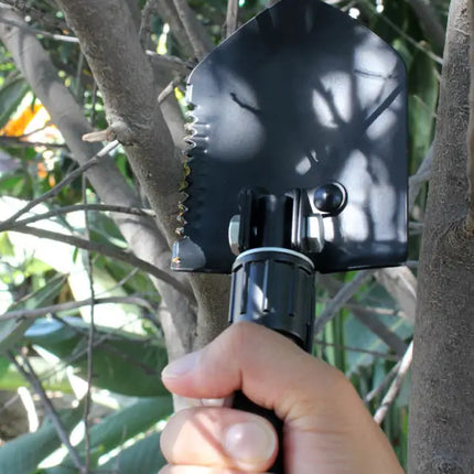 Hand holding a black multi-tool shovel with a serrated edge near tree branches