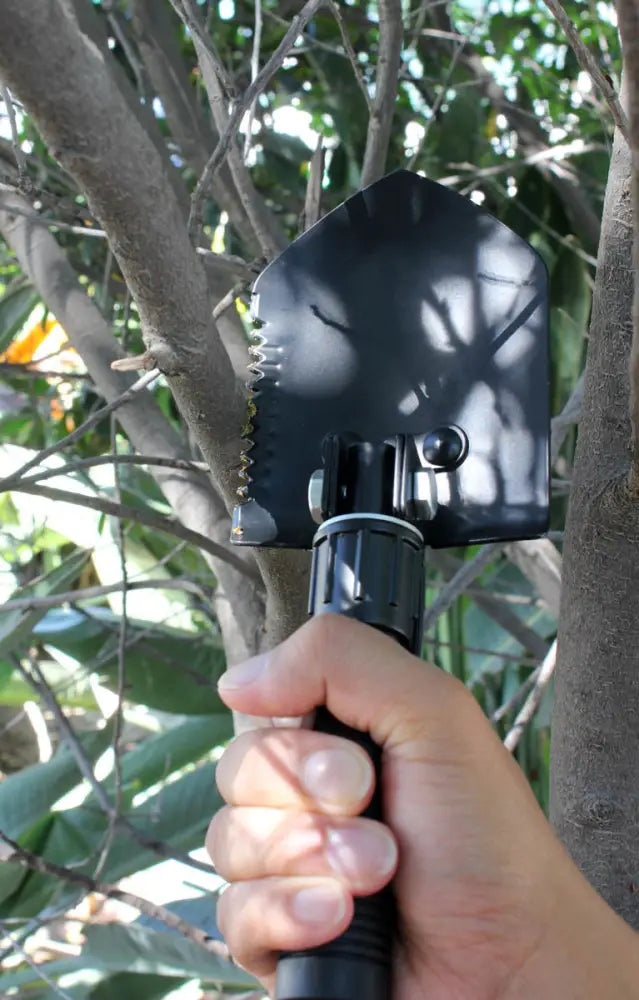 Hand holding a black multi-tool shovel with a serrated edge near tree branches