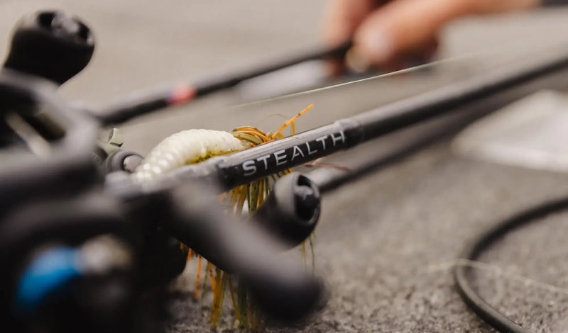 Close-up of a black fishing rod with the brand name STEALTH printed on the shaft, alongside a white and orange fly lure, with a blurred hand holding the rod in the background.