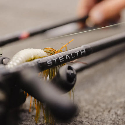 Close-up of a black fishing rod with the brand name STEALTH printed on the shaft, alongside a white and orange fly lure, with a blurred hand holding the rod in the background.