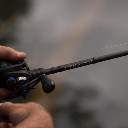 A close-up shot of a person's hands holding a black fishing rod and spinning reel, with the brand name 'STEALTH' visible on the rod, set against a blurred outdoor background.