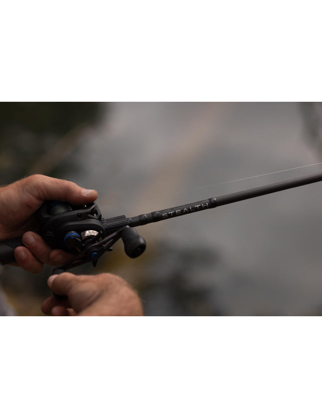 A close-up shot of a person's hands holding a black fishing rod and spinning reel, with the brand name 'STEALTH' visible on the rod, set against a blurred outdoor background.