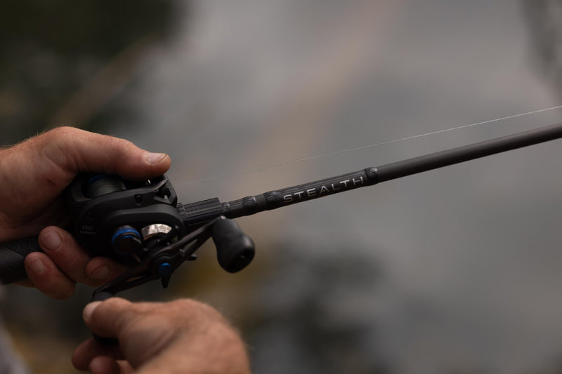 A close-up shot of a person's hands holding a black fishing rod and spinning reel, with the brand name 'STEALTH' visible on the rod, set against a blurred outdoor background.