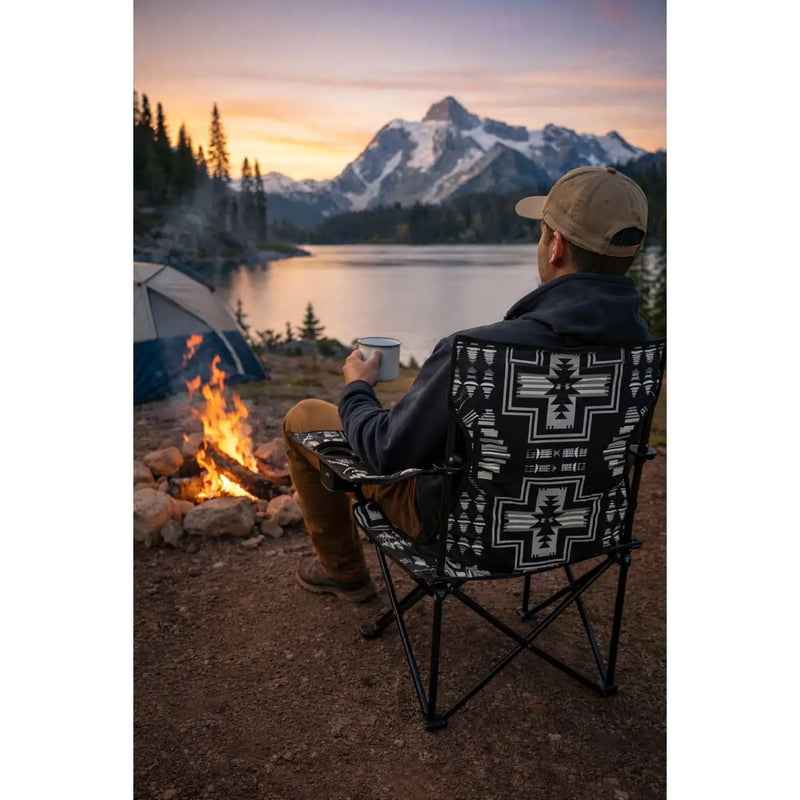 Person sitting in a patterned chair by a campfire with mountains and lake in the background
