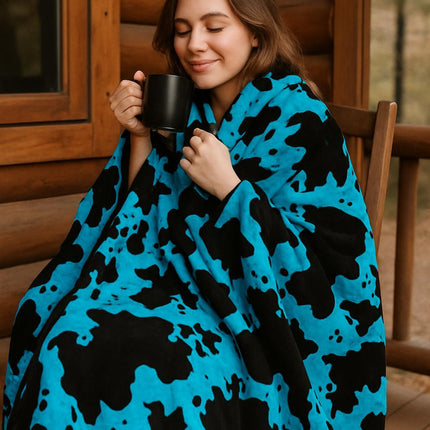 Woman wrapped in a blue and black cow patterned blanket holding a mug outdoors.