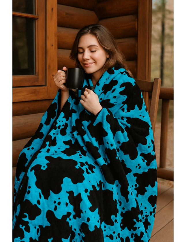 Woman wrapped in a blue and black cow patterned blanket holding a mug outdoors.