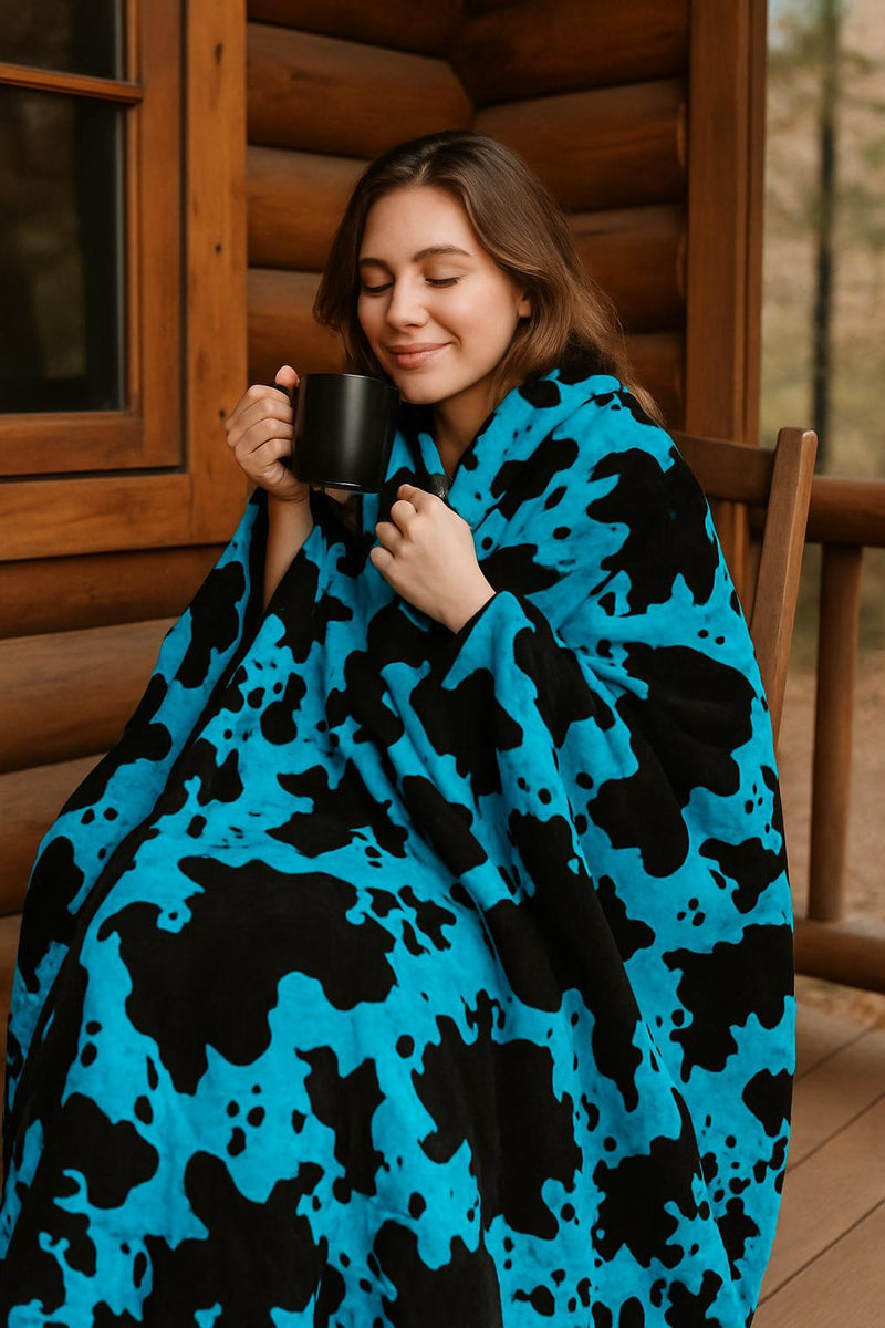 Woman wrapped in a blue and black cow patterned blanket holding a mug outdoors.