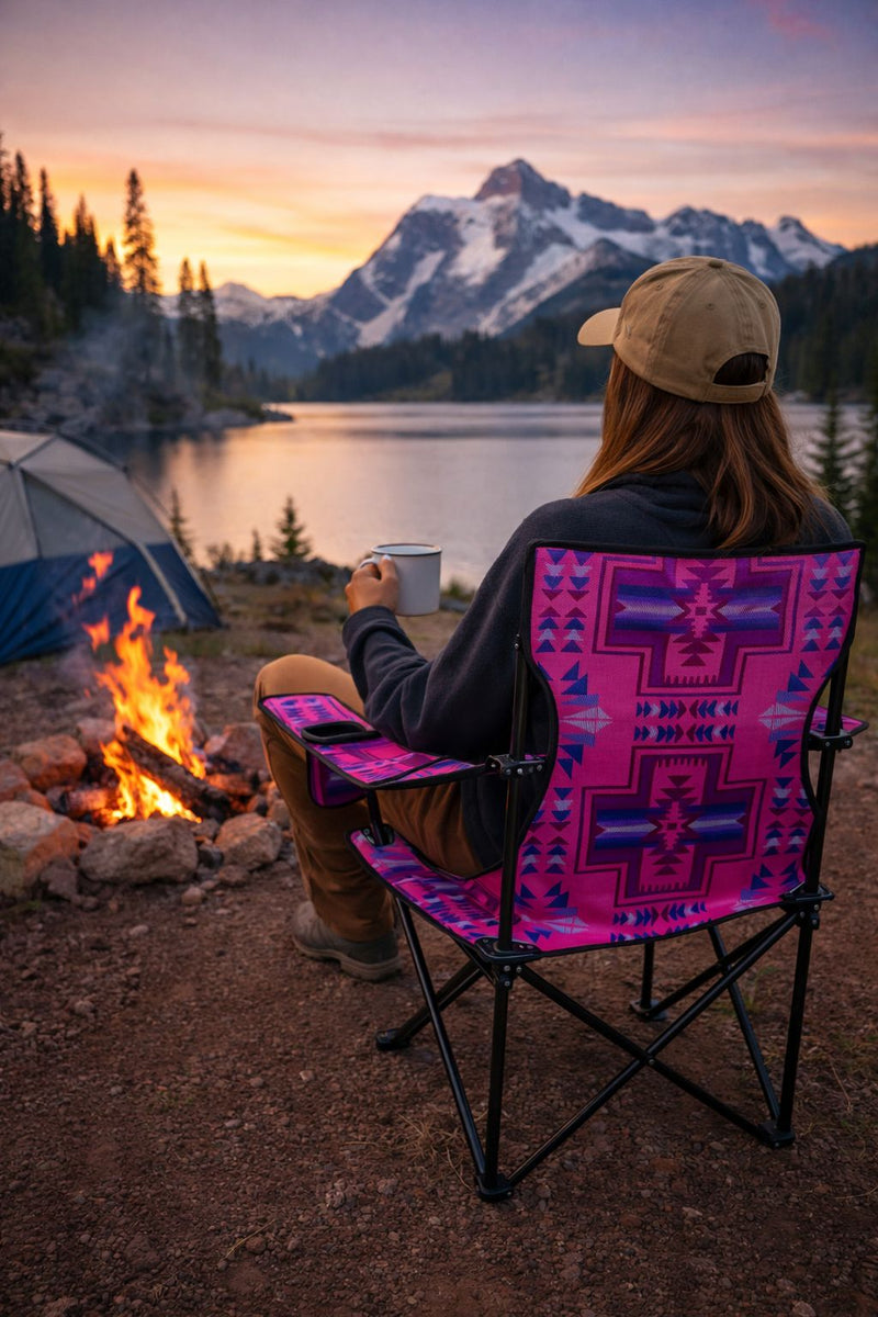Person sitting in a colorful pink, blue patterned chair by a campfire with mountains and lake in the background
