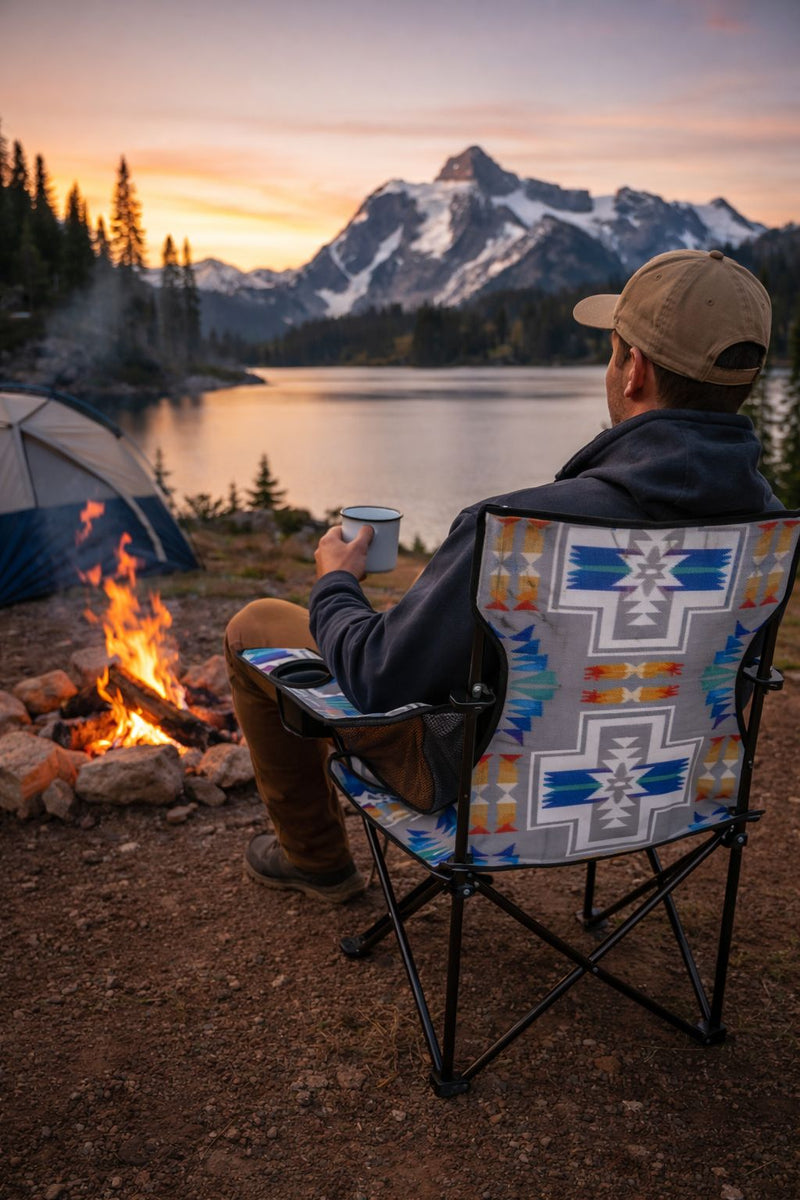 Person sitting in a grey, red, orange, blue patterned chair by a campfire with a scenic mountain view at sunset.