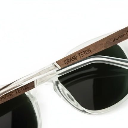 Clear frame sunglasses with dark lenses and wooden arms engraved with Grand Teton and a mountain range