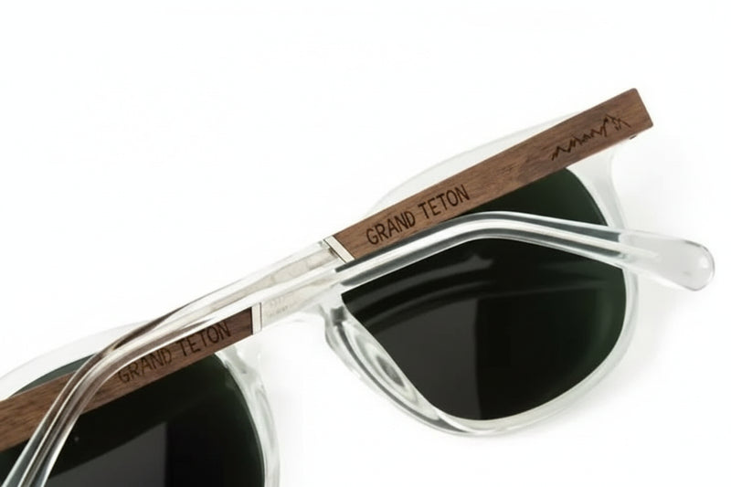Clear frame sunglasses with dark lenses and wooden arms engraved with Grand Teton and a mountain range