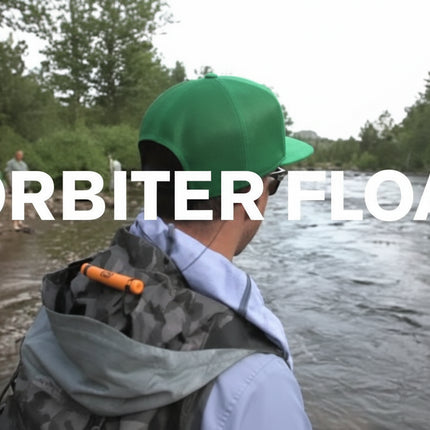 Man in green cap and grey jacket with orange item on back, looking at river with text Orbiter Float