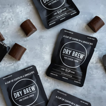 Dry Brew coffee sachets and chocolate squares scattered on a light grey surface