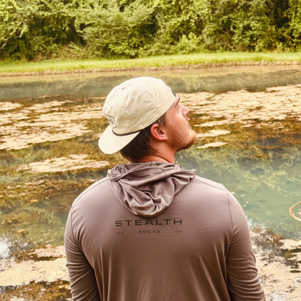 A man wearing a beige cap and a brown long-sleeve hooded shirt stands with his back to the camera, looking out over a natural landscape with water and green trees. The shirt has 'STEALTH' printed on the back.