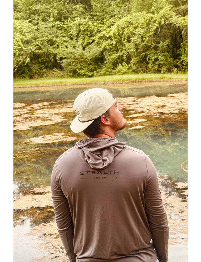 A man wearing a beige cap and a brown long-sleeve hooded shirt stands with his back to the camera, looking out over a natural landscape with water and green trees. The shirt has 'STEALTH' printed on the back.
