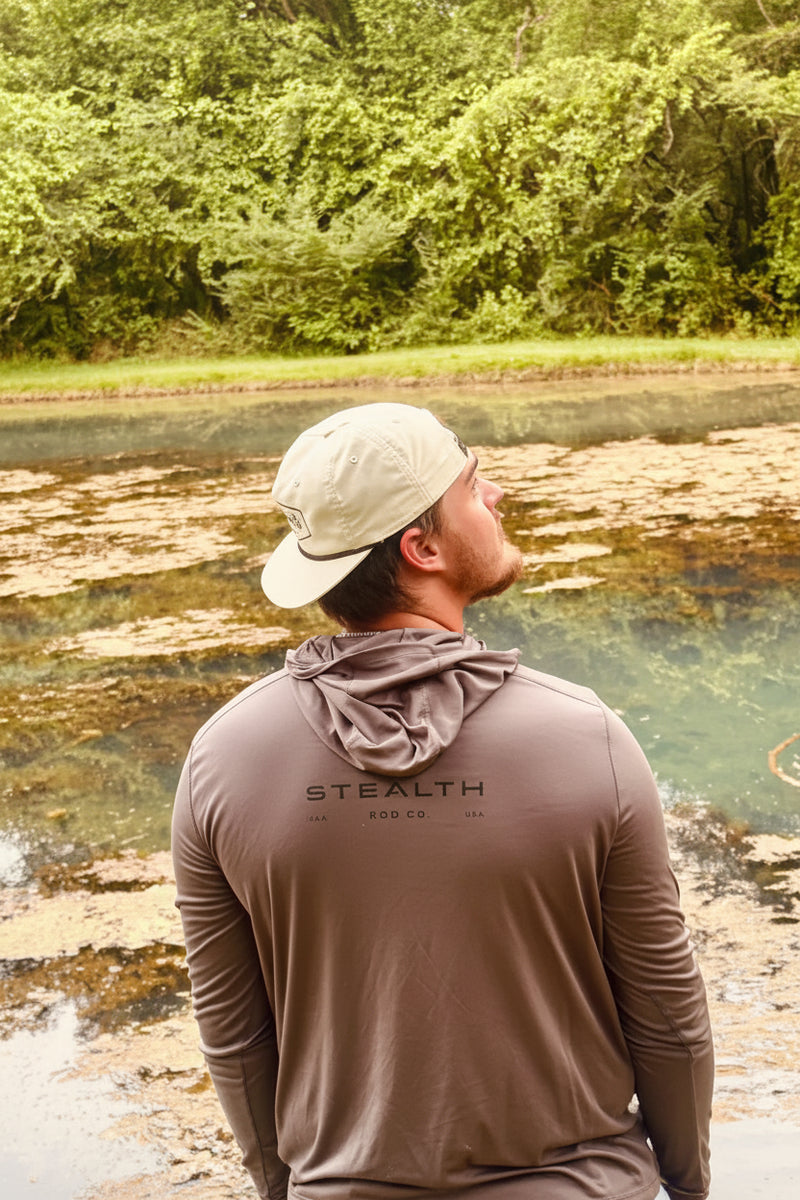 A man wearing a beige cap and a brown long-sleeve hooded shirt stands with his back to the camera, looking out over a natural landscape with water and green trees. The shirt has 'STEALTH' printed on the back.