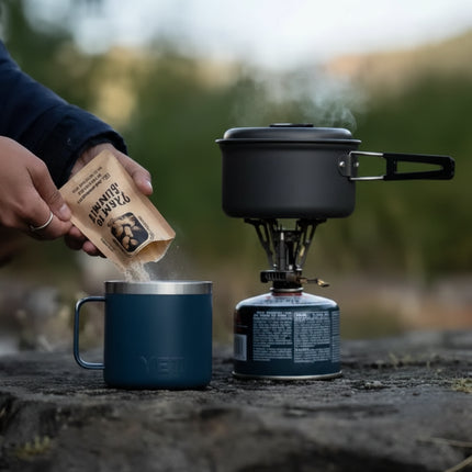 Person pouring coffee grounds into a blue mug next to a camping stove with a pot