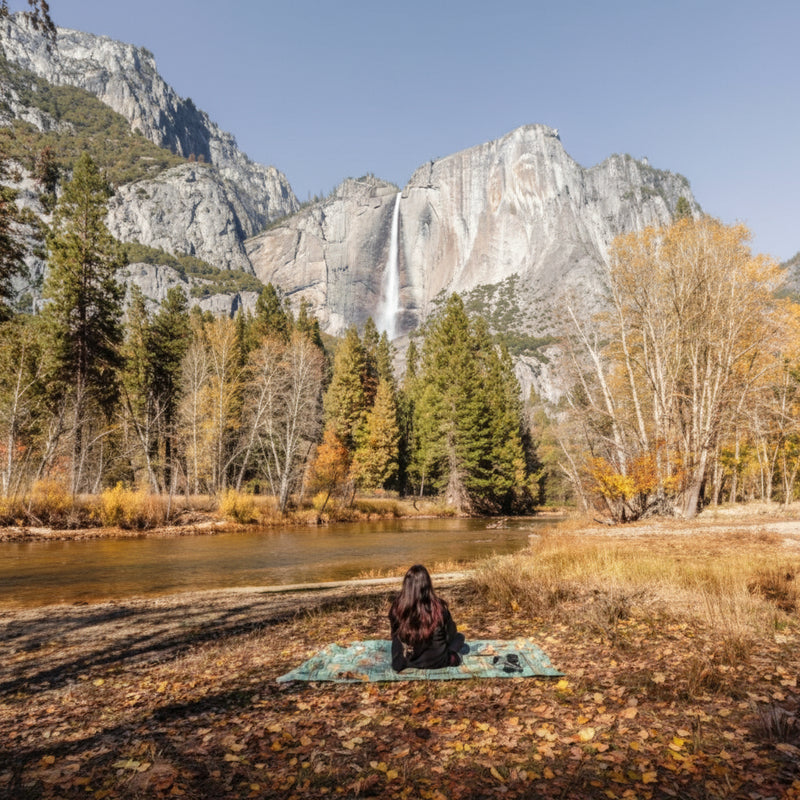 Person sitting on a mat by a river with a waterfall and mountains in the background