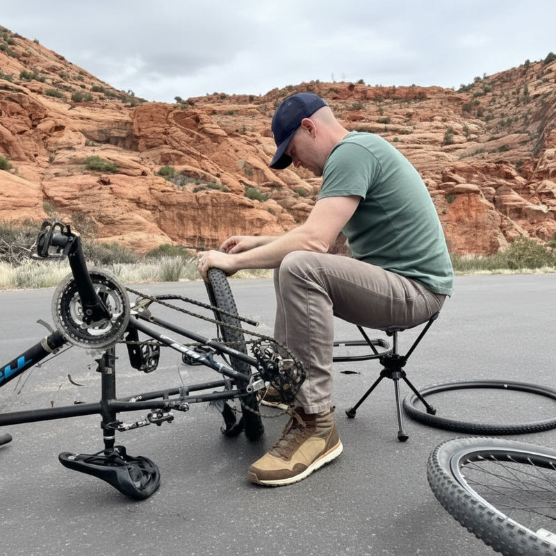 Man in a green shirt and cap repairing a bicycle on a paved road with red rock mountains in the background