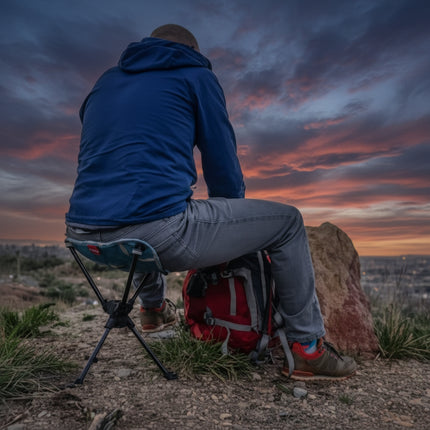Man in blue jacket and jeans sitting on a portable stool with a red backpack beside him