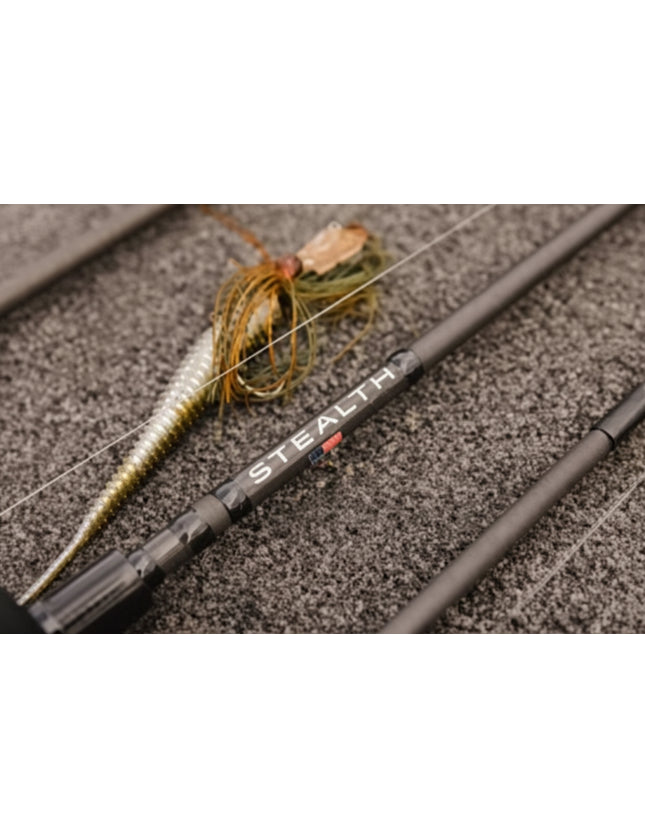 Close-up of a fishing rod with a soft plastic lure, resting on a textured gray surface. The rod features the brand name 'STEALTH' printed in white with a small red accent.
