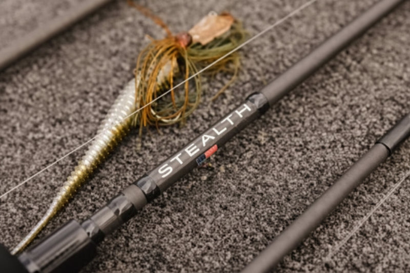 Close-up of a fishing rod with a soft plastic lure, resting on a textured gray surface. The rod features the brand name 'STEALTH' printed in white with a small red accent.