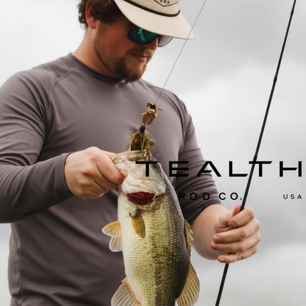 A man wearing a beige cap and sunglasses holds a largemouth bass while holding a fishing rod. The image features a dark gray overlay with the text 'STEALTH CO.