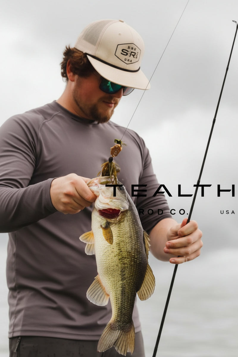 A man wearing a beige cap and sunglasses holds a largemouth bass while holding a fishing rod. The image features a dark gray overlay with the text 'STEALTH CO.