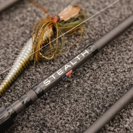 Close-up of a fishing rod with a soft plastic lure, resting on a textured gray surface. The rod features the brand name 'STEALTH' printed in white with a small red accent.