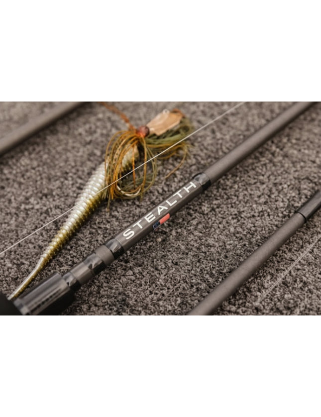 Close-up of a fishing rod with a soft plastic lure, resting on a textured gray surface. The rod features the brand name 'STEALTH' printed in white with a small red accent.