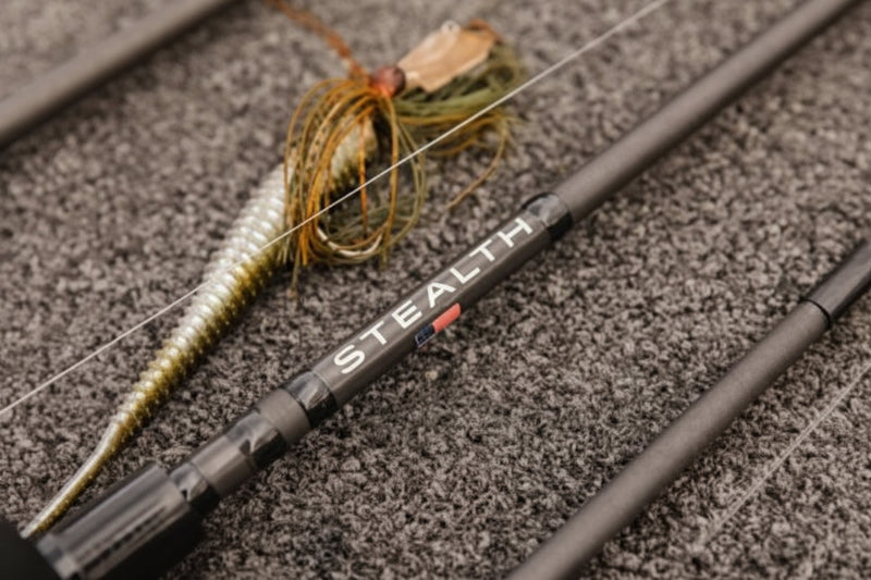 Close-up of a fishing rod with a soft plastic lure, resting on a textured gray surface. The rod features the brand name 'STEALTH' printed in white with a small red accent.