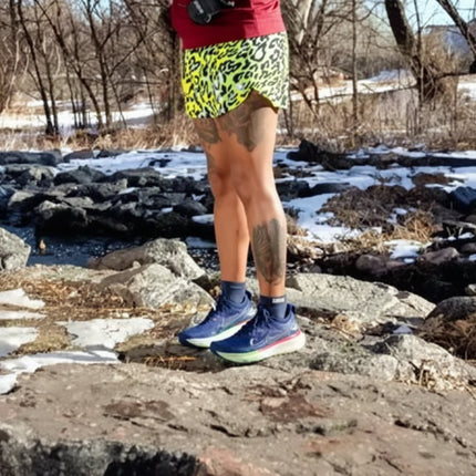 Person in yellow patterned shorts and blue running shoes standing on rocks with snow and trees with injinji toe socks ankle high.