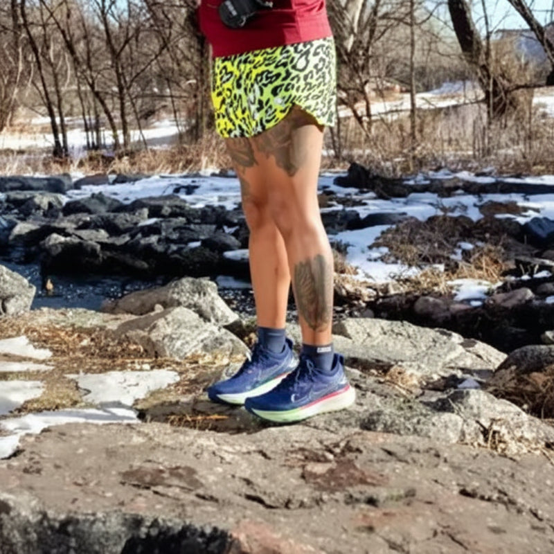 Person in yellow patterned shorts and blue running shoes standing on rocks with snow and trees with injinji toe socks ankle high.