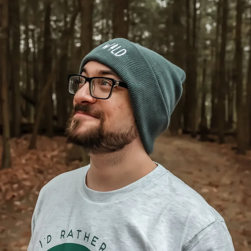 Man with beard and glasses wearing a green beanie with WILD text and a grey t-shirt in a forest